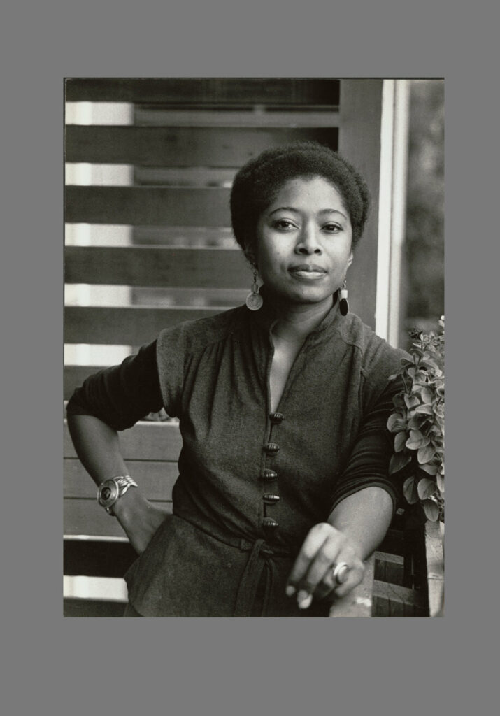 Black-and-white portrait of Alice Walker, an African-American writer and activist, standing confidently on a porch. She wears a buttoned dress with long sleeves, large round earrings, and a watch on her wrist. Her hair is styled naturally, and her expression is poised and thoughtful. A small plant is visible in the foreground to her right.