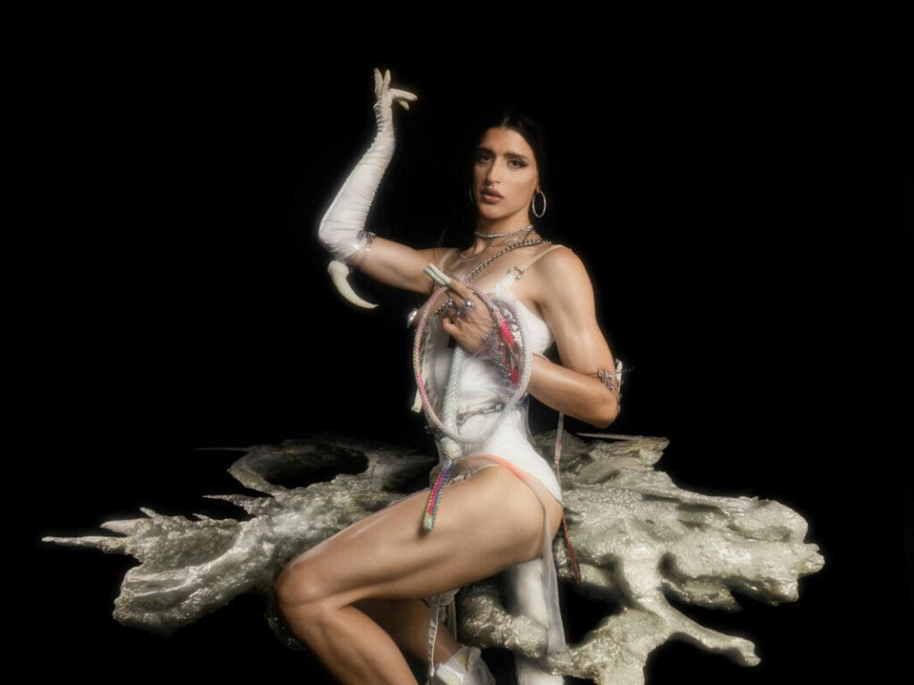 Arca sits gracefully on an abstract metallic structure resembling organic coral-like formations against a black background. She wears a sculptural white dress with asymmetric cuts, showcasing her toned arms and legs. Her ensemble includes striking long gloves with claw-like extensions and layered metallic jewelry. She gazes confidently at the viewer, her dark hair slicked back, complemented by hoop earrings. The image exudes an avant-garde and ethereal atmosphere, blending strength and elegance