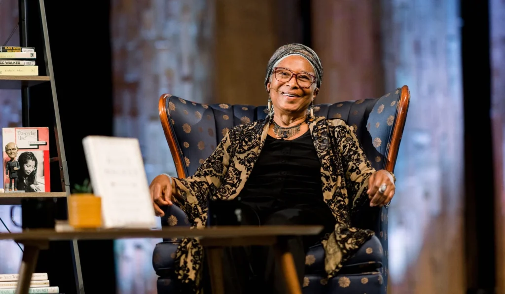 Alice Walker, an African-American writer and activist, seated comfortably in an elegant armchair on a stage. She is wearing a patterned shawl over a black outfit, a headscarf, and glasses, exuding warmth and wisdom. A bookshelf with neatly arranged books is visible to her left, and the background features a softly lit, textured design, adding depth to the setting. Her relaxed posture and gentle smile convey a welcoming and reflective presence.