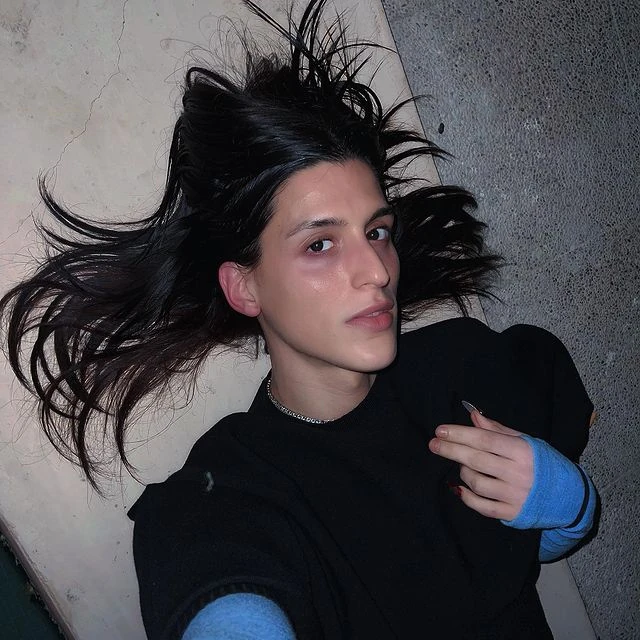 Arca lies on the ground with her dark hair fanned out in all directions, creating a striking halo effect. She wears a black top layered over light blue sleeves, adding a casual yet stylish touch. Her face is illuminated by soft lighting, highlighting her natural features, minimal makeup, and a calm, confident expression. A delicate chain necklace adds a subtle detail, while her hand is raised with two fingers partially extended, conveying a relaxed, intimate vibe. The background features a mix of textured surfaces, blending soft concrete and stone.