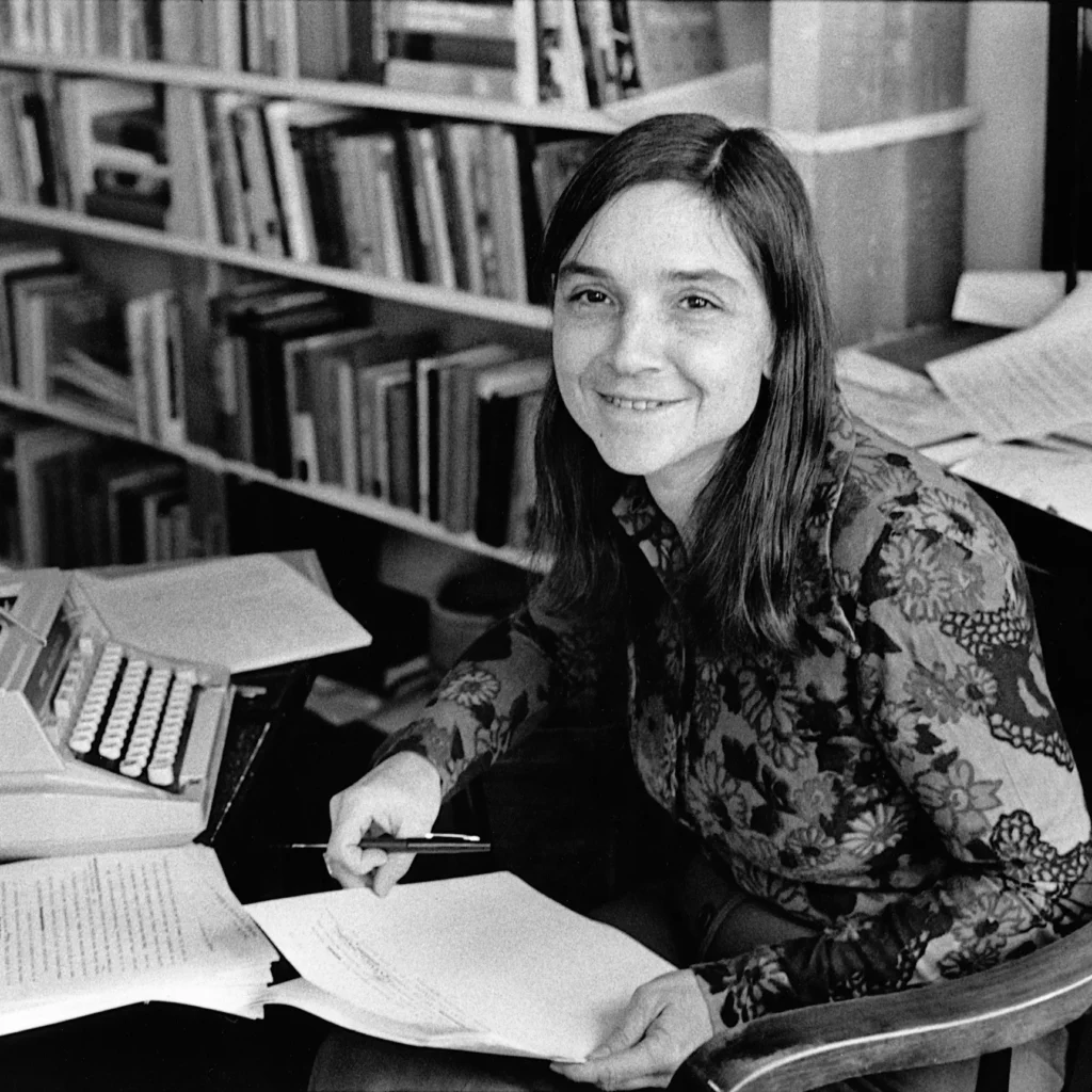 
Black-and-white photograph of Adrienne Rich sitting at a desk with papers in hand and a typewriter to her side. She is wearing a patterned blouse and smiling softly. Bookshelves filled with books are visible in the background, highlighting her literary environment.