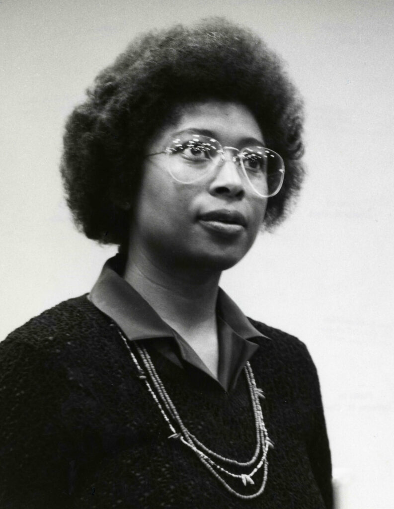 Black-and-white portrait of Alice Walker, an African-American writer and activist, wearing large round glasses and a layered necklace. She has a natural afro hairstyle and is dressed in a textured sweater over a collared shirt. Her expression is composed and thoughtful, with a neutral background emphasizing her dignified presence.