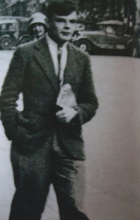 A young Alan Turing walking outdoors, dressed in a suit and tie, holding a small object or book in one hand while his other hand is in his pocket. The black-and-white photograph shows a street in the background with vintage cars and a person wearing a helmet visible, capturing a candid moment of Turing’s life in an urban setting.