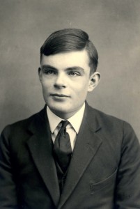 A black-and-white portrait of Alan Turing as a young man, dressed formally in a suit and tie. His hair is neatly combed, and he looks slightly off-camera with a calm and composed expression. The photograph reflects his youth.