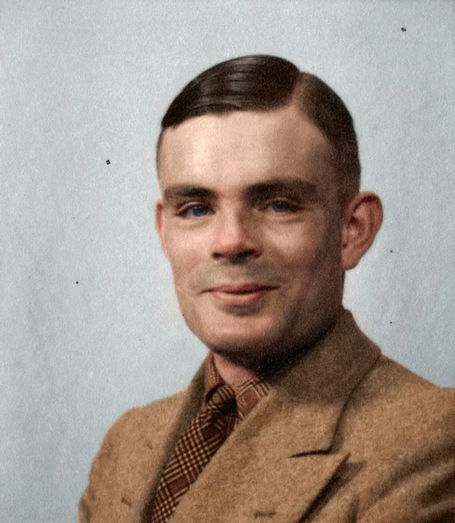 A colorized photograph of Alan Turing as an adult, wearing a light brown suit jacket and a checkered tie. His hair is neatly combed to the side, and he is smiling softly.