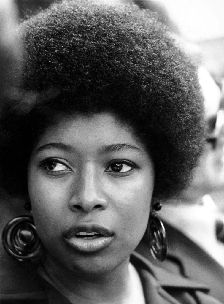 Black-and-white photograph of a young Alice Walker, an African-American writer and activist, with a voluminous afro hairstyle. She wears bold, circular earrings and has a focused, expressive gaze, her mouth slightly open as if in mid-speech or thought. The background is softly blurred, highlighting her striking features and presence.