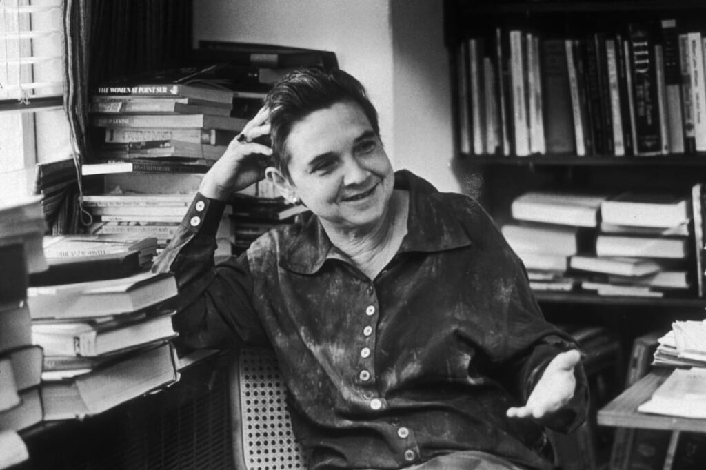 Black-and-white photograph of Adrienne Rich sitting in a room filled with books. She is leaning her head on one hand, smiling gently, and wearing a button-up shirt. Bookshelves and stacks of books surround her, reflecting her scholarly and literary life.