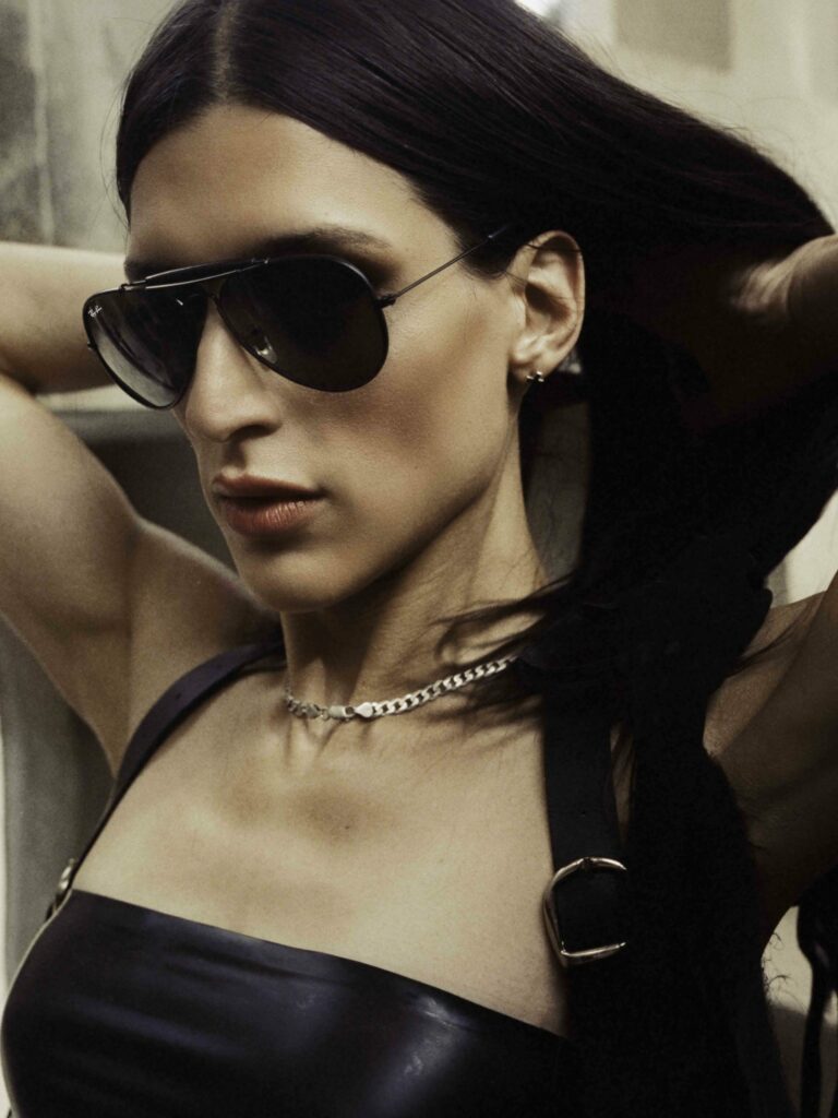 Arca adjusts her long, dark hair while wearing sleek, dark aviator sunglasses that exude a bold and commanding presence. She is dressed in a fitted black leather outfit with silver buckle details on the straps, adding a sharp and edgy touch to her look. A delicate chain necklace adorns her neck, complementing her ensemble. Her expression is intense and focused, framed by a softly blurred background, emphasizing her dynamic and modern aesthetic.