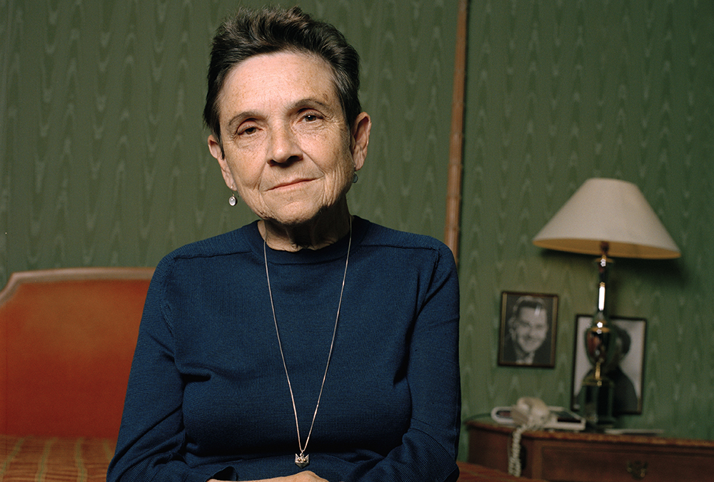 Color photograph of Adrienne Rich seated in a room with a green patterned wall and a lamp on a bedside table behind her. She is wearing a dark blue top and a simple necklace, her short hair styled neatly. Framed photos and personal items are visible on the table, adding a sense of intimacy and warmth to the setting.