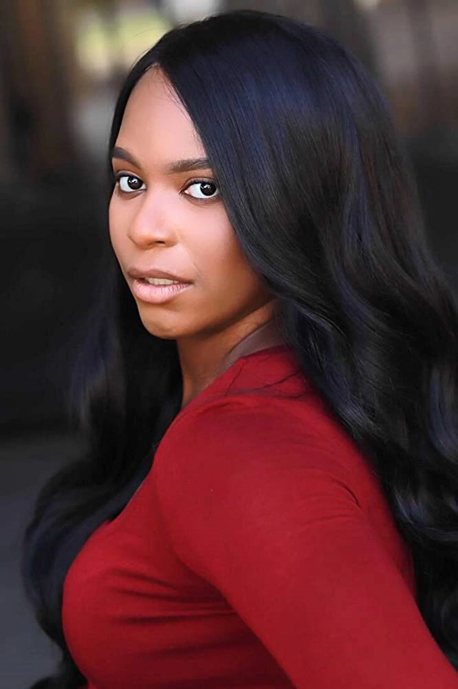 Alexandra Grey stands outdoors, wearing a fitted red top that contrasts beautifully with her long, glossy black hair. She looks back over her shoulder with a piercing, confident gaze. The blurred background enhances the focus on her poised expression and elegant presence.