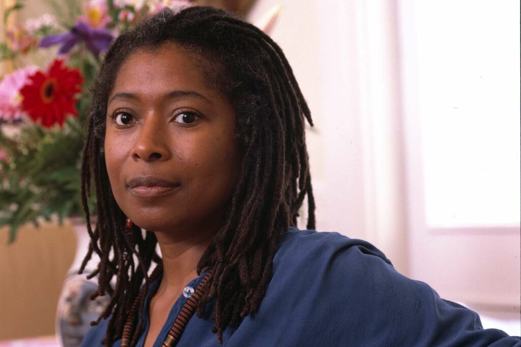 Alice Walker, an African-American writer and activist, sits in a brightly lit room, wearing a deep blue blouse. Her long dreadlocks fall naturally over her shoulders, and she gazes directly at the camera with a calm and composed expression. In the background, a vase of colorful flowers adds vibrancy to the setting, complementing the serene and inviting atmosphere of the portrait.