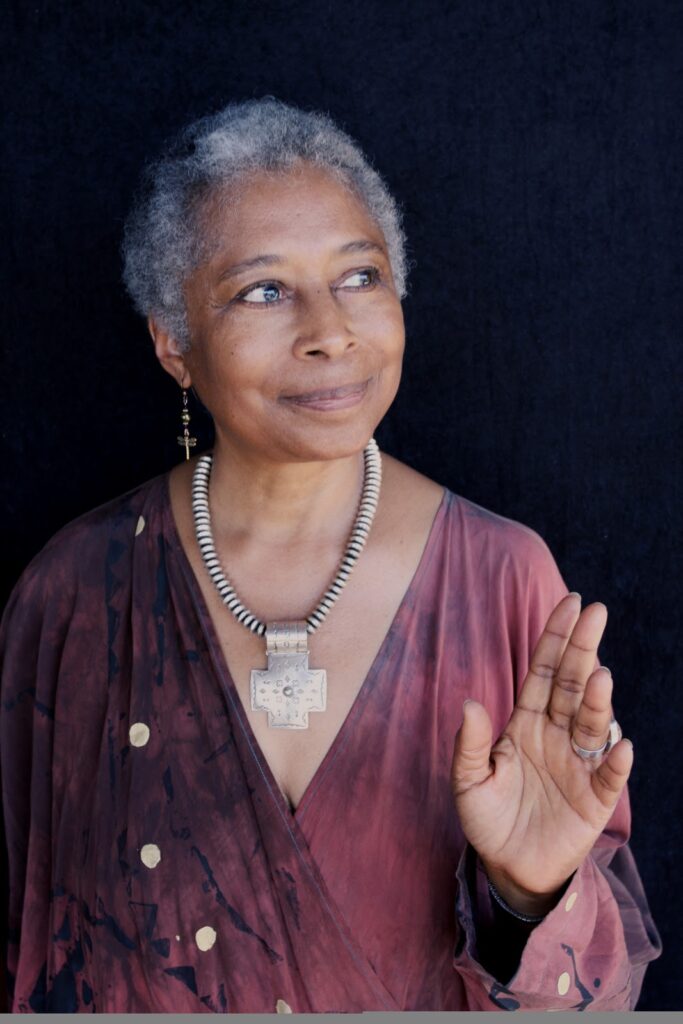 Alice Walker, an African-American writer and activist, stands against a dark background wearing a flowing, patterned garment in earthy tones. She accessorizes with a striking necklace featuring a large cross pendant and a pair of earrings, one shaped as a cross. Her short gray hair complements her serene expression, as she gently raises her hand in a gesture of calm or acknowledgment. The overall image conveys grace, wisdom, and spiritual presence.