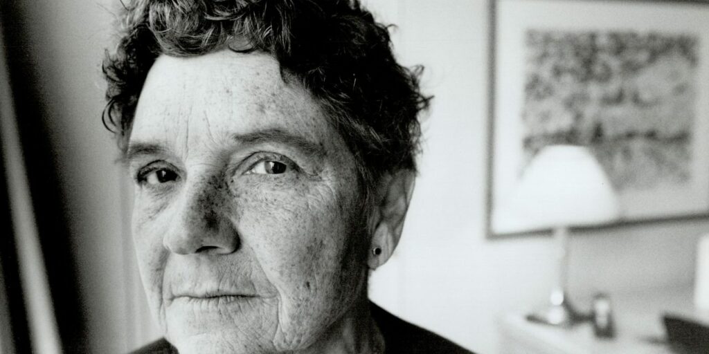 Black-and-white close-up photograph of Adrienne Rich with short, curly hair and a thoughtful expression. The background features a softly blurred room with a lamp and framed artwork, giving the image a personal and reflective atmosphere.