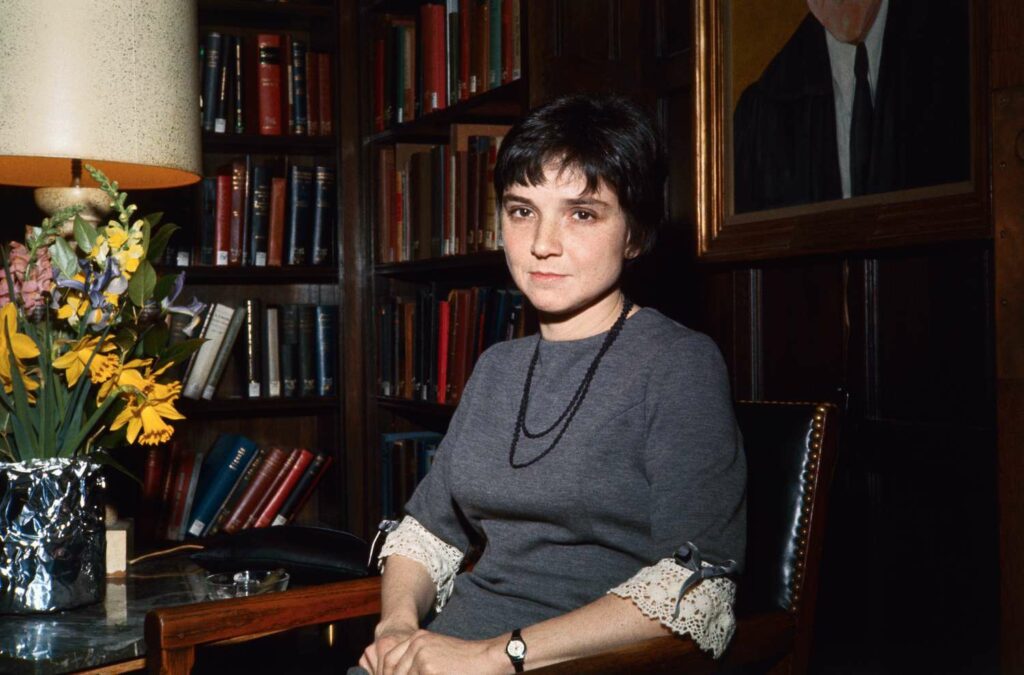 Color photograph of a young Adrienne Rich seated in a room filled with bookshelves and a table adorned with a vase of flowers. She is wearing a grey dress with lace-trimmed sleeves and a simple necklace, gazing calmly at the camera. The background includes a lamp and a framed portrait, emphasizing a scholarly and elegant setting.