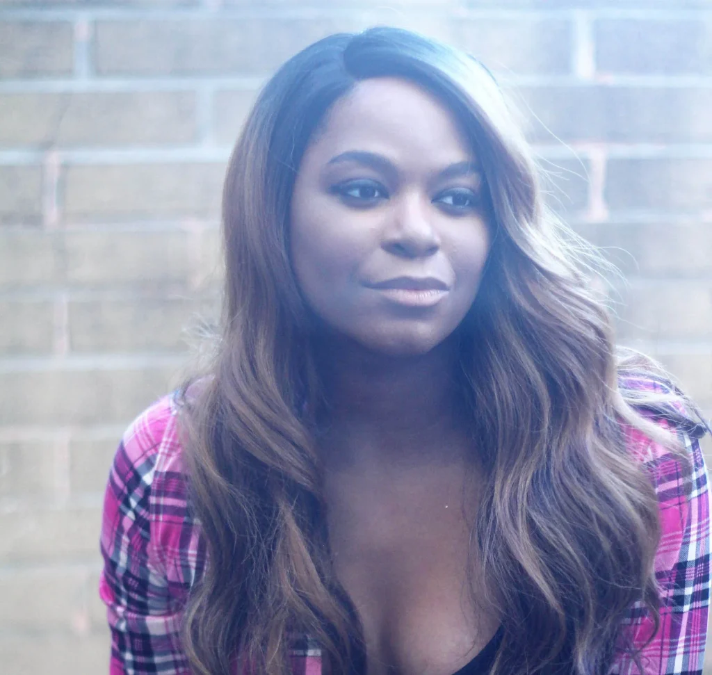 Alexandra Grey is pictured in a relaxed moment against a softly lit brick wall background. She wears a pink plaid shirt over a black top, with her long, wavy brown hair cascading over her shoulders. Her calm expression and the gentle lighting create a warm, contemplative atmosphere.