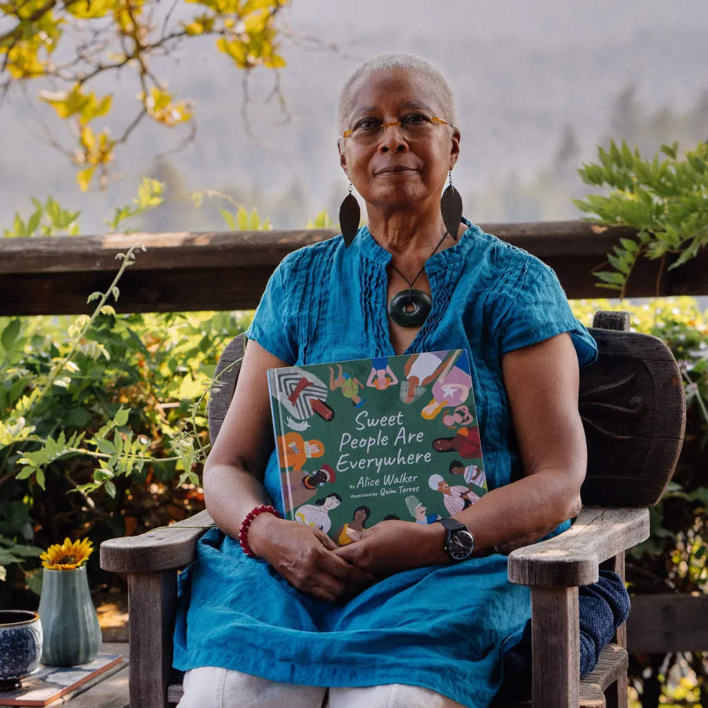 Alice Walker, an African-American writer and activist, sits outdoors on a wooden bench, holding her book Sweet People Are Everywhere. She is wearing a vibrant blue top, large statement earrings, and glasses. Her calm and thoughtful expression matches the serene natural setting, surrounded by greenery and soft sunlight. A small vase with a yellow flower is visible on a table to her side, adding warmth to the peaceful scene.