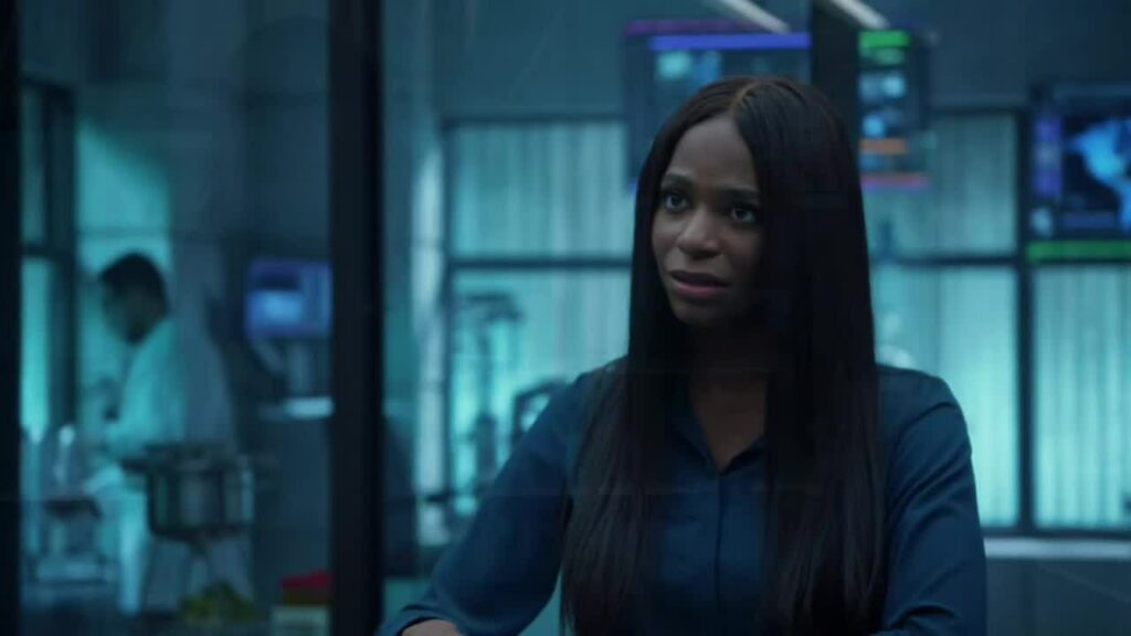 Grey appears in a laboratory setting, wearing a dark blue top. The background features blurred scientific equipment, monitors, and another person in a lab coat working. Her expression is thoughtful and focused, reflecting a professional or intense moment in the scene. The environment suggests a medical or scientific context.