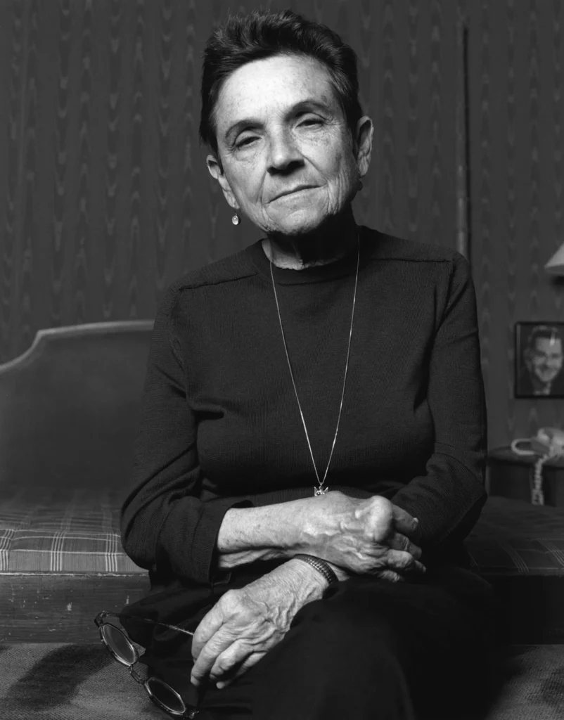 Black-and-white photograph of Adrienne Rich seated on a bed, her hands resting gently in her lap. She is wearing a dark top and a long necklace, gazing directly at the camera with a calm and reflective expression. The background features a patterned wall and framed photographs, adding a sense of personal and intimate atmosphere.