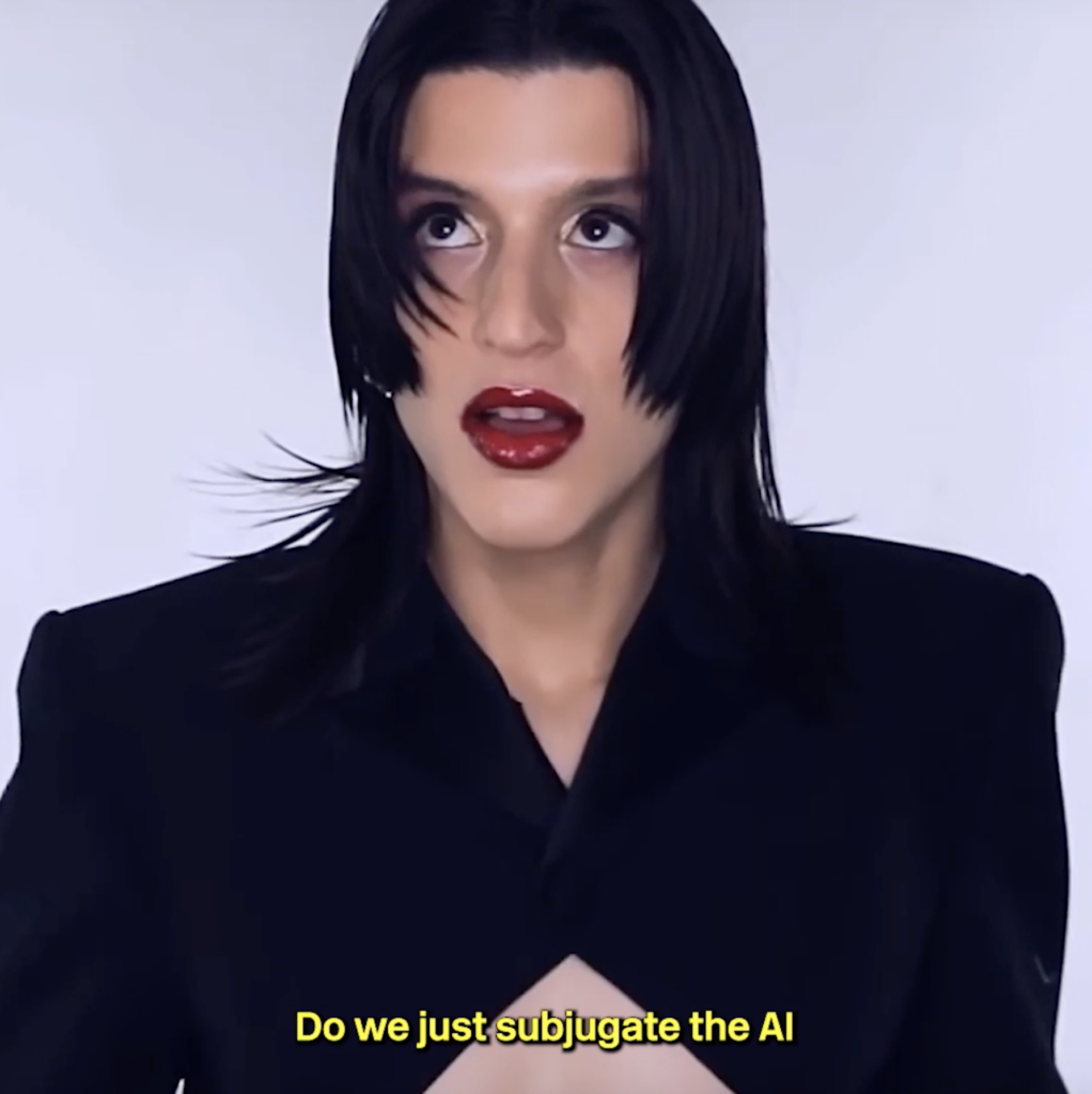 Arca is captured mid-expression, looking upward with wide eyes and parted lips painted in bold red lipstick. She wears a sharp black blazer with structured shoulders, exuding a dramatic and thought-provoking presence. The image features subtitles at the bottom that read, "Do we just subjugate the AI," adding an intellectual and futuristic layer to the composition. The plain white background enhances the focus on her striking features and the philosophical undertone of the moment.