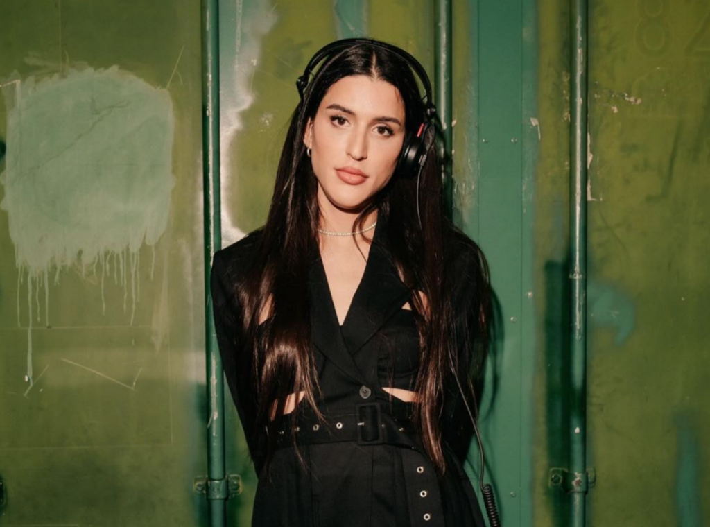 Arca stands confidently against a textured green wall, wearing a sleek black outfit with cut-out details and a cinched belt, exuding a modern and stylish vibe. She has long, dark, flowing hair and is wearing headphones, adding a subtle nod to her musical artistry. Her expression is composed and inviting, with soft makeup emphasizing her features. The industrial-style background contrasts with her polished look, highlighting her dynamic presence and individuality.