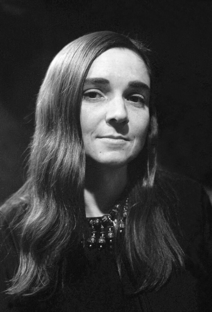 A black-and-white portrait of Adrienne Rich in her younger years. She has long, smooth hair parted slightly off-center and wears a dark top with an intricate beaded neckline. Her expression is thoughtful and composed as she gazes softly at the camera, the lighting highlighting her features against a dark background.