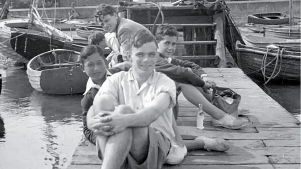 A young Alan Turing, smiling and seated on a dock with his legs crossed, wearing shorts and a casual shirt. He is accompanied by three friends, all relaxed and enjoying a moment by the water. Small boats are docked nearby, contributing to a serene and cheerful atmosphere, hinting at Turing's lighter, more personal moments away.