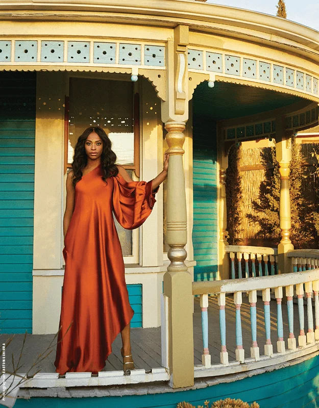 Alexandra Grey stands gracefully on the porch of a vibrant, Victorian-style house painted in teal and cream. She wears a flowing, one-shoulder burnt-orange gown with dramatic sleeve details, exuding elegance and poise. The golden sunlight casts a warm glow, enhancing the rich tones of her dress and the architectural details of the house. Her confident stance and serene expression embody strength and sophistication.