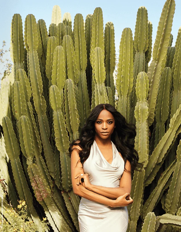 Alexandra Grey stands confidently in front of a towering cactus backdrop, wearing a sleek, elegant white halter dress. Her long black hair flows over her shoulders as she crosses her arms, exuding grace and composure. The natural setting contrasts beautifully with her sophisticated appearance, creating a striking and harmonious image.