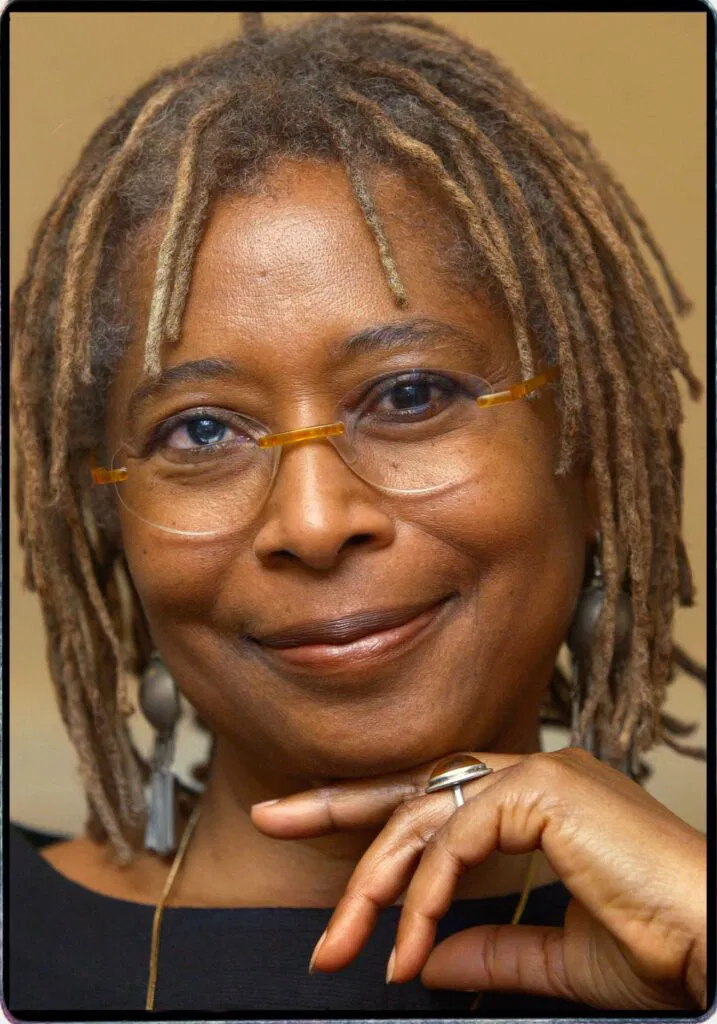 Alice Walker, an African-American writer and activist, smiles gently in this close-up portrait. She has shoulder-length dreadlocks with natural highlights and wears glasses with thin frames. Her hand rests thoughtfully near her chin, adorned with a ring. She is dressed in a simple black top and long earrings, exuding warmth, intelligence, and confidence against a neutral background.