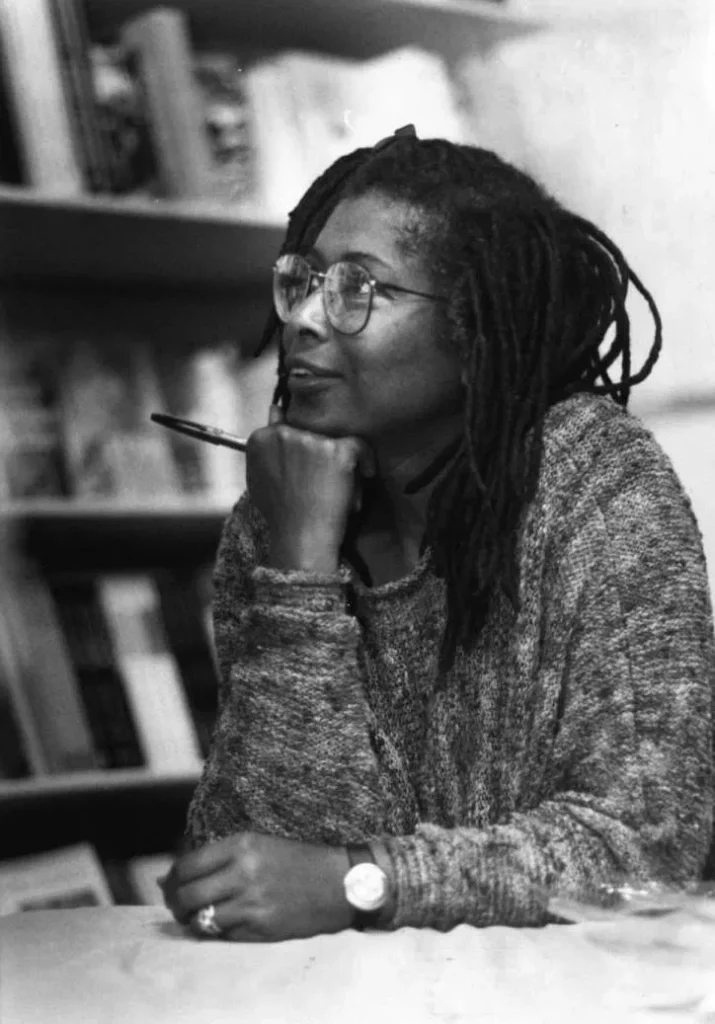 Black-and-white photograph of Alice Walker, an African-American writer and activist, seated at a table in a contemplative pose. She rests her chin on her hand, holding a pen, while wearing glasses and a cozy sweater. Her long dreadlocks frame her face. Shelves filled with books are visible in the blurred background, emphasizing her connection to literature and intellectual thought. The image captures a moment of quiet reflection and creativity.