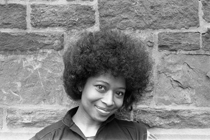 Black-and-white portrait of a young Alice Walker, an African-American writer and activist, standing in front of a textured stone wall. She has a radiant smile and wears her natural afro hairstyle with pride. Dressed in a simple top, her expression exudes warmth, confidence, and a hint of playfulness. The rustic background contrasts beautifully with her vibrant presence.