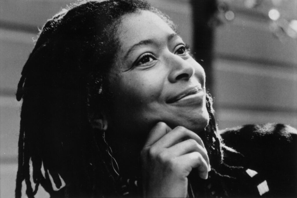 Black-and-white close-up portrait of Alice Walker, a prominent African-American writer and activist. She gazes upward with a serene and thoughtful expression, her chin resting gently on her hand. Her long dreadlocks frame her face, and the soft background highlights her contemplative demeanor. The image exudes a sense of calm and introspection.