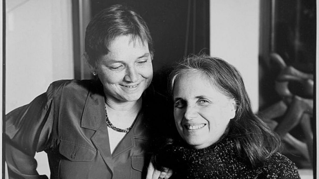 Black-and-white photograph of Adrienne Rich smiling warmly while standing beside another woman, likely her partner Michelle Cliff. Rich is wearing a collared shirt and a necklace, while the other woman is dressed in a textured sweater. The image captures an intimate and joyful moment between the two, with a blurred background adding focus to their expressions.