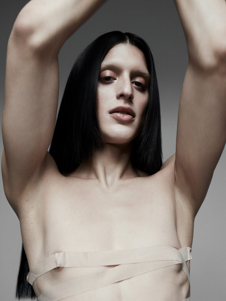 Arca stands with her arms raised above her head, creating an elongated and graceful silhouette. Her long, straight black hair cascades down her shoulders, contrasting with her pale skin. She wears a minimalist nude-toned harness that subtly wraps around her chest, blending with her skin tone and adding an avant-garde element. Her expression is soft yet confident, embodying both vulnerability and strength. The neutral gray background enhances the focus on her striking features and minimalist aesthetic.