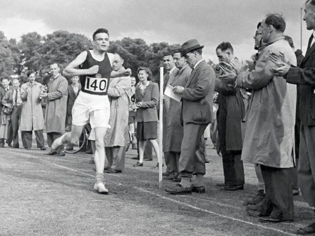 Alan Turing crossing the finish line during a race in 1946, wearing a running bib numbered 140. He is dressed in athletic shorts and a sleeveless top, exuding determination. Spectators and officials, some with clipboards, line the grassy track, observing and applauding the moment. The scene highlights Turing's passion for running and his dedication beyond his intellectual pursuits.