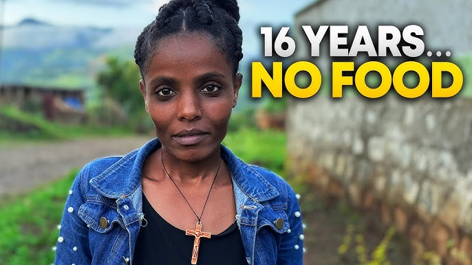 This image features Muluwork, the Ethiopian woman known for her extraordinary claim of surviving without food or water for 16 years. She is depicted standing outdoors, wearing a denim jacket and a cross necklace, with a determined expression. The background showcases a rural setting with greenery and a stone wall. Overlaid text reads, "16 YEARS... NO FOOD," emphasizing the remarkable nature of her story.