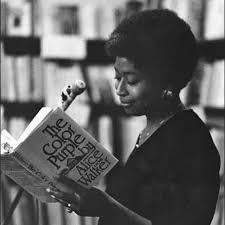 Black-and-white photograph of Alice Walker reading from her novel The Color Purple at a public event. She stands in front of a microphone, holding the book open, with shelves of books blurred in the background. Her short afro and elegant demeanor reflect her focus and poise as she shares her work. The scene captures a moment of literary engagement and connection.