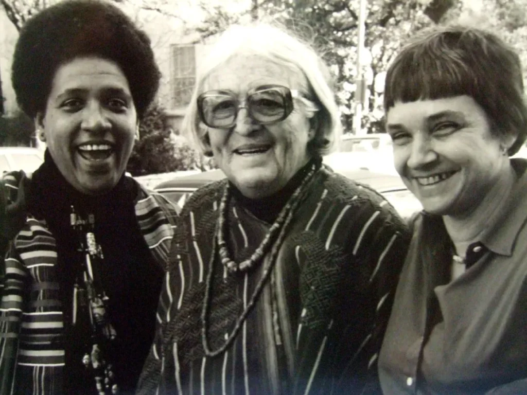 Black-and-white photograph of Audre Lorde, Meridel Le Sueur, and Adrienne Rich standing outdoors, smiling warmly. Lorde is on the left, wearing a patterned jacket and a necklace, Le Sueur is in the center with glasses and beads, and Rich is on the right in a collared shirt. The trio exudes camaraderie and shared purpose against a backdrop of trees and parked cars.