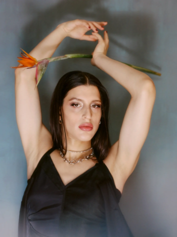 Arca poses gracefully with her arms raised above her head, holding a single vibrant orange tropical flower with a long green stem. She wears a flowing black dress with thin straps, exuding a soft, ethereal elegance. A delicate chain necklace adorns her neck, while her dark, sleek hair frames her face. Her expression is serene yet confident, with a subtle gloss on her lips. The background is a muted gradient of blue and gray, enhancing the dreamlike quality of the composition.