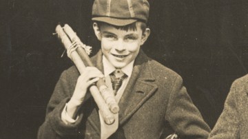 A young Alan Turing, dressed in a school uniform and cap, smiles warmly while holding a bundle of sticks over his shoulder. The sepia-toned photograph captures him during his school years, radiating a sense of youthful curiosity and energy.