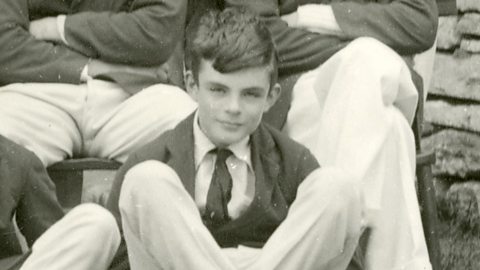 A young Alan Turing sitting casually on the ground with his legs crossed, wearing a blazer, tie, and light-colored trousers. He gazes directly at the camera with a slight smile. Behind him, other young men in formal attire are seated on stone steps, suggesting a school or group setting.