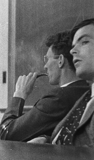 A candid black-and-white photograph of Alan Turing sitting in a classroom or lecture setting. Turing is in the foreground, wearing a suit and patterned tie, slightly turned with a thoughtful expression. In the background, another man, wearing glasses and holding a pipe, appears to be focused on the discussion. A chalkboard is visible, suggesting an academic environment.