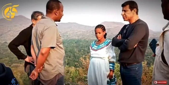 This image features Muluwork, standing among a group of individuals during an outdoor meeting. She is dressed in a white traditional Ethiopian dress with colorful accents, appearing engaged in conversation. The setting includes a scenic backdrop of rolling hills and lush greenery, emphasizing the rural environment where she resides. The group’s body language suggests a discussion or investigation, aligning with the interest in Muluwork's extraordinary claim of living without food or water for 16 years.