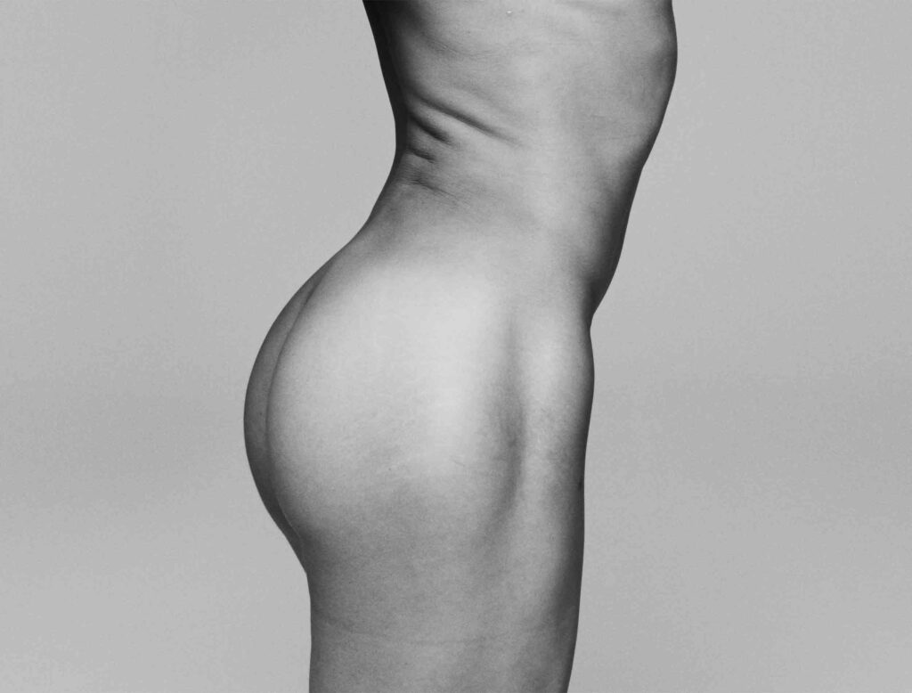 A monochromatic side profile of Arca's torso and lower body, showcasing her form in a minimalist and artistic manner. The smooth contours and lighting emphasize the natural curves and textures of her body against a plain gray background. The image highlights themes of self-expression, body positivity, and the fluidity of identity in a bold yet understated presentation.