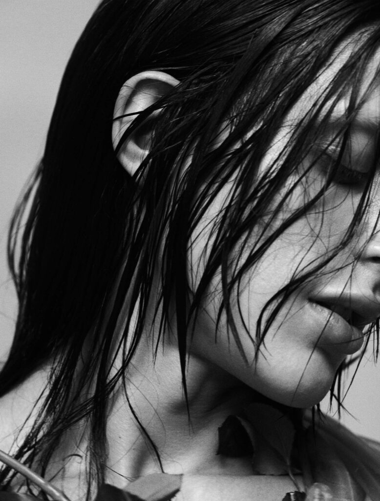 A close-up black-and-white portrait of Arca, her wet hair cascading over her face in an evocative and raw composition. Her eyes are closed, and her slightly parted lips suggest introspection and vulnerability. The textured lighting accentuates the natural contours of her face and neck, while the abstract details of her outfit add an ethereal quality. The minimalist background ensures full focus on the emotional intensity of the image.