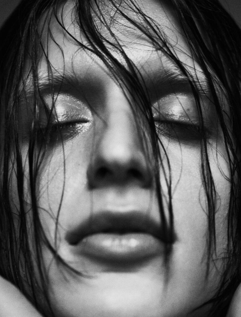 An intimate black-and-white close-up of Arca's face, with wet strands of hair framing her features. Her eyes are softly closed, and her slightly parted lips convey a sense of vulnerability and serenity. The subtle shimmer on her eyelids adds a delicate contrast to the raw, natural texture of the image. The tight framing and minimal background focus attention on the emotional depth and intensity of her expression.