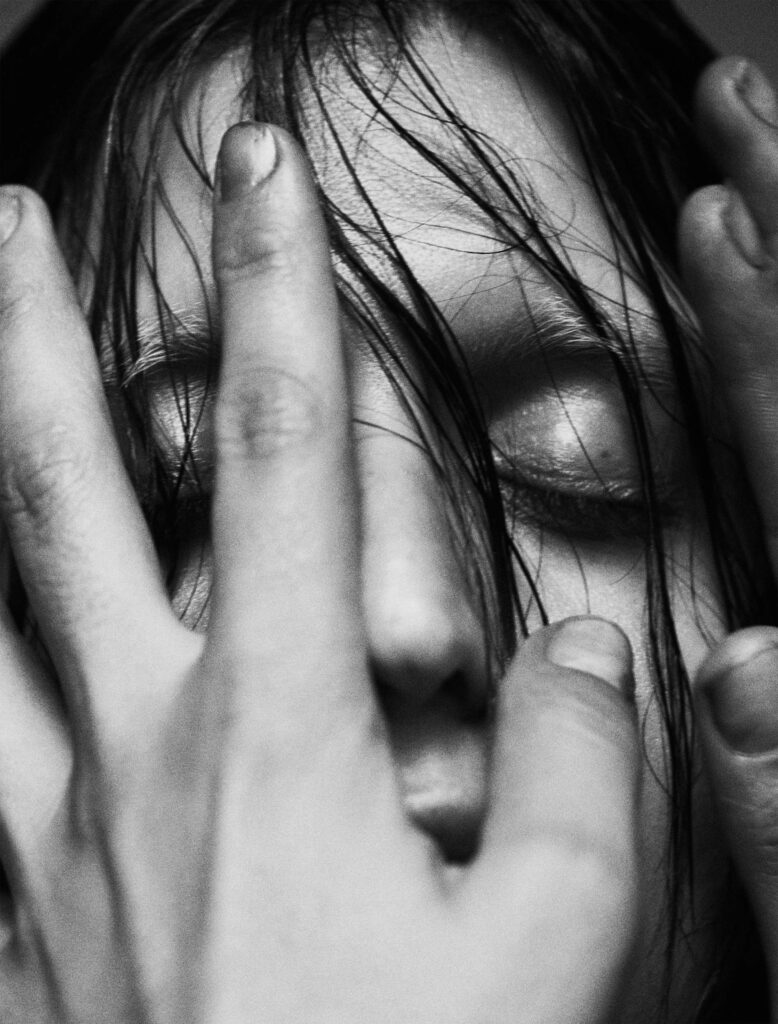 A black-and-white close-up of Arca’s face, partially obscured by her hands and wet strands of hair. Her eyes are closed, creating a serene and introspective mood, while her fingers gently frame her face in an expressive, almost sculptural pose. The contrast between the soft lighting and the raw textures of her skin and hair adds depth and emotion to the image. The minimalist background focuses attention on her enigmatic presence.