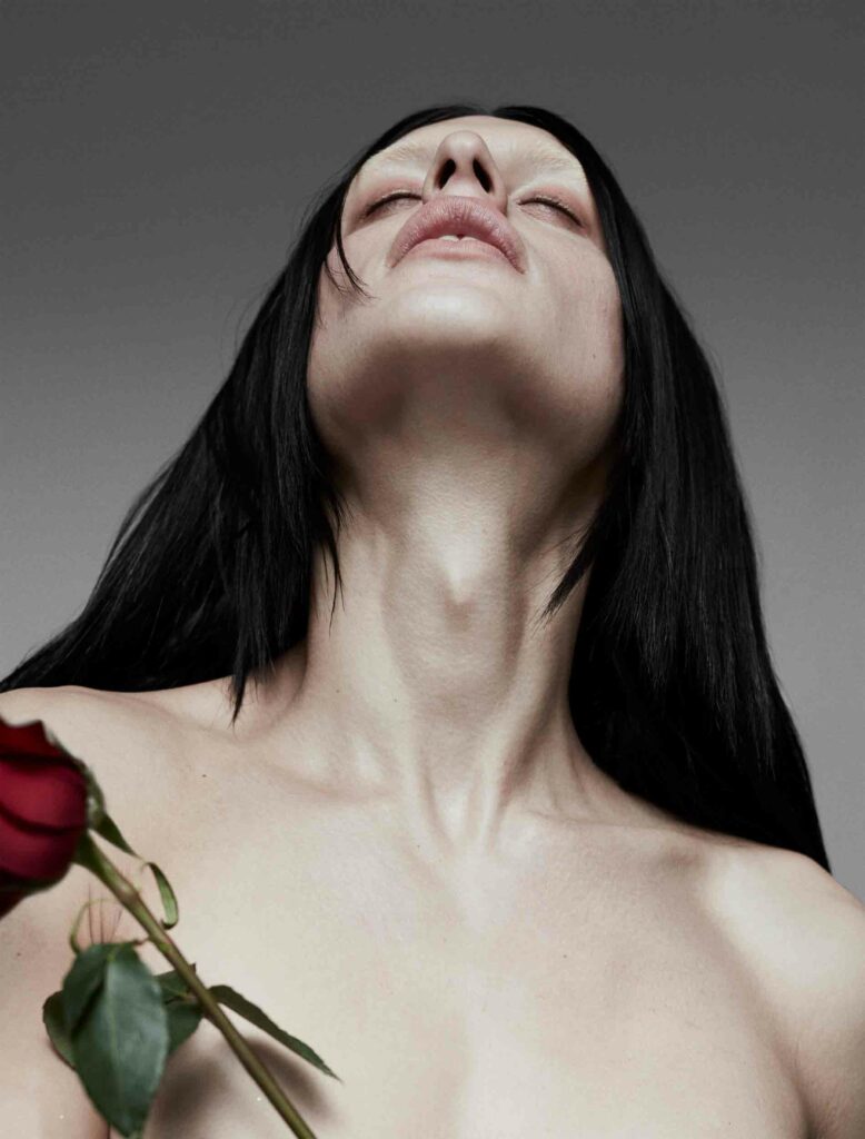 A dramatic portrait of Arca with her head tilted back, her face upward, exposing the elegant curve of her neck. Her long black hair cascades down her shoulders, and she holds a single red rose with visible green leaves in the foreground. The muted gray background emphasizes the contrast between her pale skin and the vibrant rose, creating a blend of vulnerability and strength in the composition. The pose evokes a sense of surrender or transcendence, highlighting her ethereal presence.