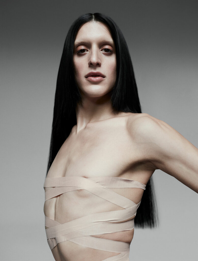 A striking portrait of Arca with long, sleek black hair cascading over her shoulders, standing confidently against a neutral gray background. Her torso is wrapped in pale, crisscrossed fabric, accentuating her angular features and toned physique. Her direct gaze into the camera conveys a blend of poise and intensity, embodying both vulnerability and power. The minimalist composition emphasizes her androgynous beauty and avant-garde aesthetic.