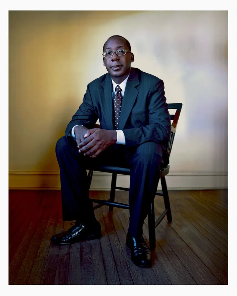 Cornelius Baker sits on a wooden chair in a formal portrait. He wears a dark suit with a patterned tie and glasses. The background has a soft gradient of warm tones, and he looks directly at the camera with a composed expression.